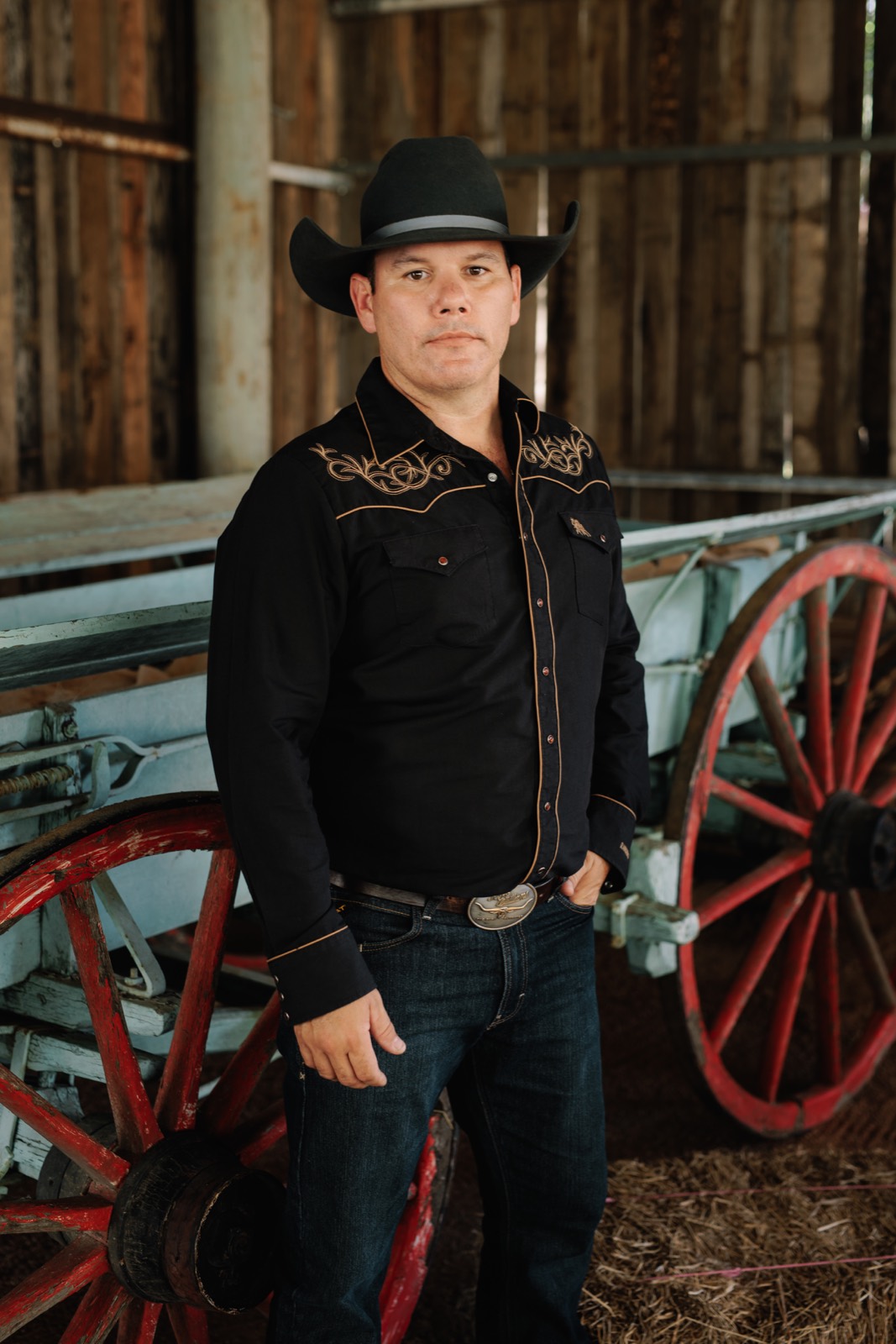 Luke Geiger in a black hat and embroidered western shirt, standing by an old timber wagon